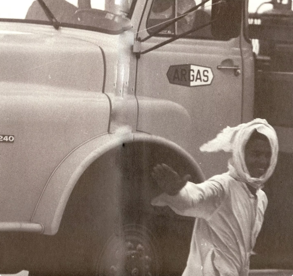 Historical black-and-white photo featuring a man waving beside a vintage truck displaying the historic ARGAS logo.