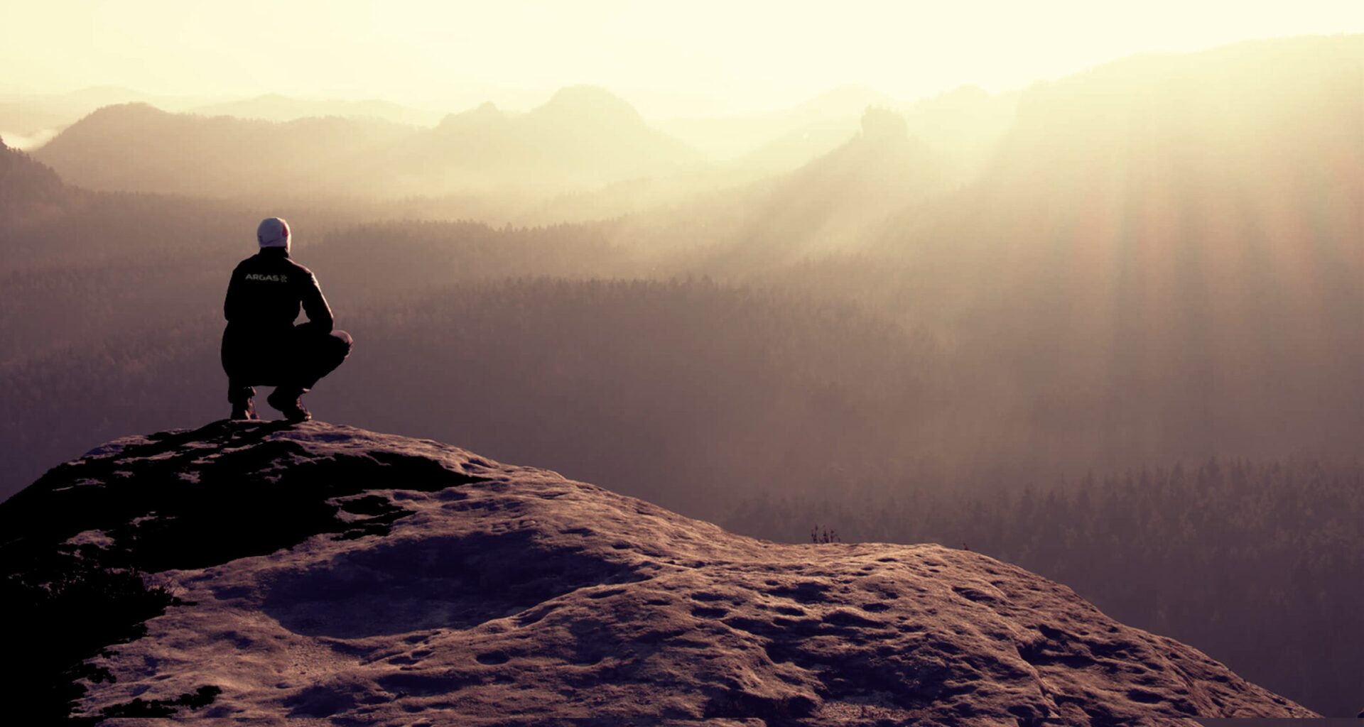 An ARGAS employee stands on a rock, gazing at the hazy mountains in the distance, with warm tones dominating the scene.